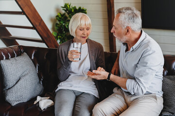 Elderly man giving to his sick wife with glass of water pills at home. Husband taked care wife...