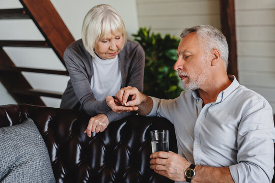 Elderly woman giving her sick husband a glass of water with pills at home. Wife cares for his health. Senior couple, man taking medicine or vitamin supplements for treatment.