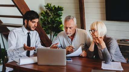 Smiling senior couple sit at lawyer broker designer office look at laptop listen to offer proposal...