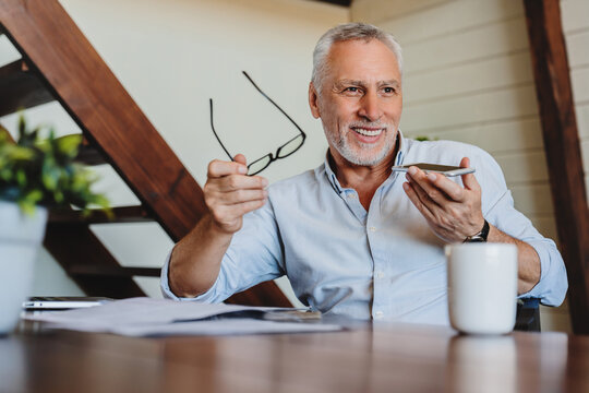 Senior caucasian man using voice search application on smartphone sitting at laptop in modern office. Male entrepreneur talking by cellphone in loud speaker mode at workplace