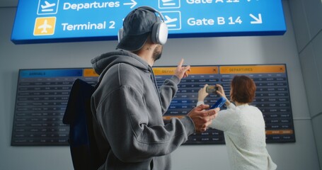 Busy Airport Terminal: Man with Backpack Checks Flight Information on Mobile Phone, Looks at Digital Arrival and Departure Board. Traveler Going on Holiday Trip. Diverse People Walking In Background.