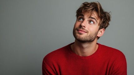 Obraz premium Surprised young man with tousled hair wearing a vibrant red sweater looking up with a smile against a soft gray studio background.