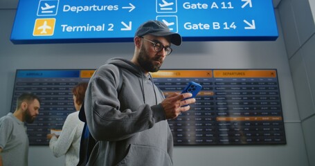 Airport Terminal: Man with Backpack Checks Flight Information Scanning Ticket Using Phone, Looks at...