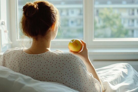 A young woman in a light polka-dotted shirt holds an apple while gazing thoughtfully out of a sunlit window. Recovery and Nutrition, Vitality Amid Illness
