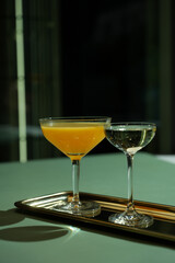 A two cocktails on a tray. One cocktail is orange and the other is clear in martini glass