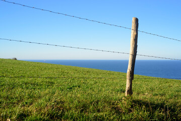 Fototapeta premium Valla de alambres y piquetes de madera en pradera de hierba verde junto al horizonte marino en Asturias
