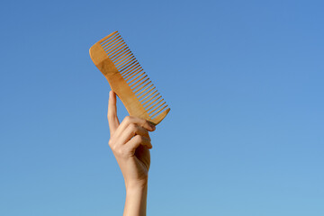 Naklejka premium Female hand holding natural wooden comb on blue sky background. Hair combing and hygiene concept, natural eco hair products