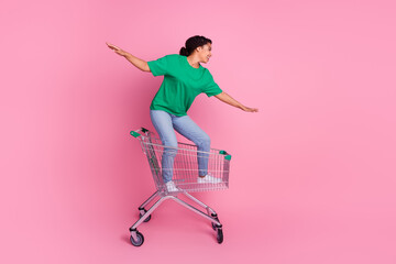 Full length photo of lovely young lady ride shopping cart have fun balance dressed stylish green garment isolated on pink color background © deagreez