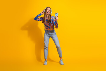 Fototapeta premium Stylish young woman enjoying music on headphones and holding smartphone, standing against vibrant yellow background