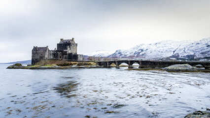 Eilean Donan Castle with bridge under a grey clouded sky and snowy mountains in the background in Scotland
