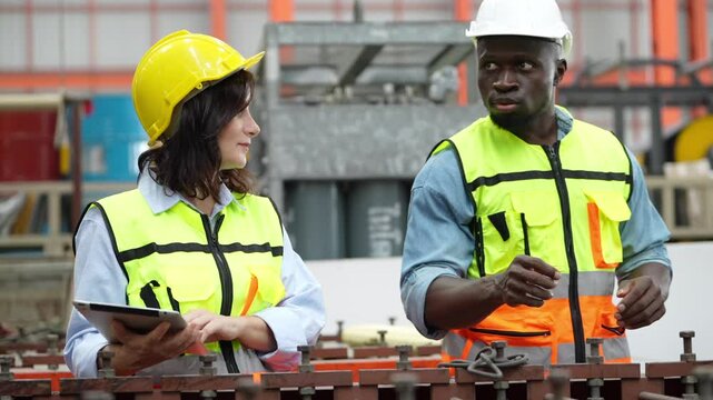 Industrial Collaboration: Two construction workers in hard hats and safety vests discuss plans with a tablet computer inside a factory setting.