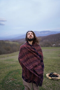 Full length portrait of bearded man wrapped in ethnic woven blanket standing outdoors on hills