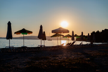 Serene beach sunset with umbrellas and sailboat on calm water by the shore in the evening