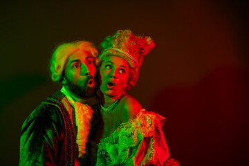 Elegant couple dressed in classic masquerade costumes illuminated by colorful light against a dark...