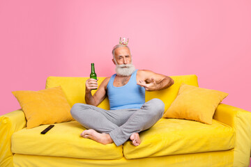 Senior man relaxing indoors wearing a crown, seated on a yellow sofa with a beer bottle, pink...