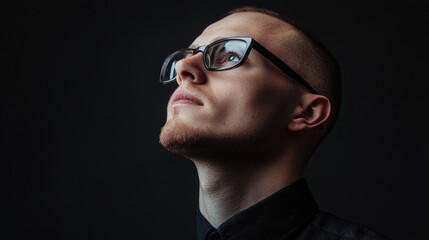 Obraz premium Contemplative Young Man with Glasses Gazing Upwards Against Dark Background in Studio Environment, Expressive Portrait with Subtle Lighting
