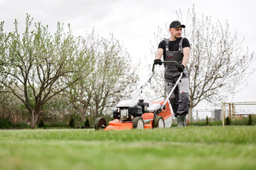Professional gardener in protective apparel is mowing green grass lawn using modern gasoline...
