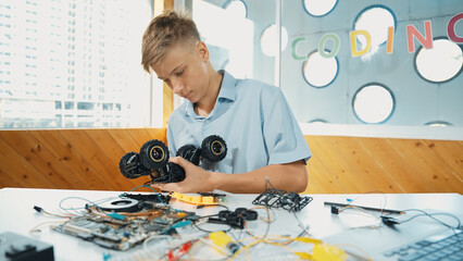 Highschool boy fixing car model with laptop and electric tool placed on table. Closeup of teenager inspect robotic machine construction while using eletronic equipment in STEM classroom. Edification.