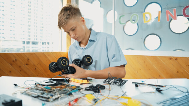 Highschool boy fixing car model with laptop and electric tool placed on table. Closeup of teenager inspect robotic machine construction while using eletronic equipment in STEM classroom. Edification.