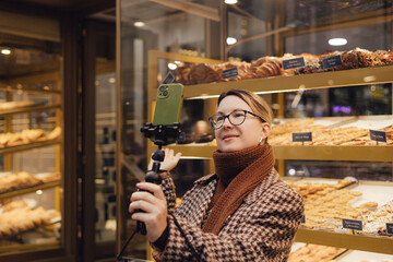 Middle age traveler woman recording video on phone with traditional Portuguese desserts Pastel de nata in the window of a bakery in Lisbon. UGC selfie