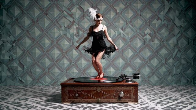 Young female dancer wearing 1920s flapper dress and headpiece dancing charleston on top of a vintage wooden gramophone record player in a room with retro wallpaper