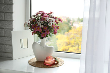 Stylish vase with beautiful flowers and burning candle near window at home
