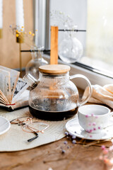 espresso coffee in a white cup on a white saucer on a wooden tray under the sun's rays, a transparent teapot with coffee, flowers, a book, morning coffee, drink, light composition, still life