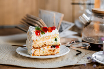 a piece of cake with buttercream on pieces of multi-colored jelly on a white saucer on a wooden tray under the sun's rays, espresso coffee in a white cup, a transparent teapot with coffee, flowers