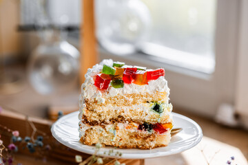 a piece of cake with buttercream on pieces of multi-colored jelly on a white saucer on a wooden tray under the sun's rays, espresso coffee in a white cup, a transparent teapot with coffee, flowers