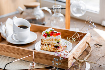 a piece of cake with buttercream on pieces of multi-colored jelly on a white saucer on a wooden tray under the sun's rays, espresso coffee in a white cup, a transparent teapot with coffee, flowers