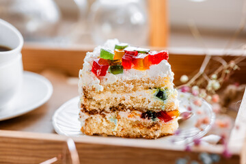 a piece of cake with buttercream on pieces of multi-colored jelly on a white saucer on a wooden tray under the sun's rays, espresso coffee in a white cup, a transparent teapot with coffee, flowers