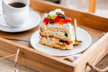 a piece of cake with buttercream on pieces of multi-colored jelly on a white saucer on a wooden tray under the sun's rays, espresso coffee in a white cup, a transparent teapot with coffee, flowers