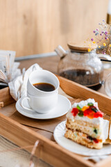 a piece of cake with buttercream on pieces of multi-colored jelly on a white saucer on a wooden tray under the sun's rays, espresso coffee in a white cup, a transparent teapot with coffee, flowers