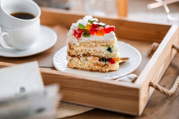 a piece of cake with buttercream on pieces of multi-colored jelly on a white saucer on a wooden tray under the sun's rays, espresso coffee in a white cup, a transparent teapot with coffee, flowers