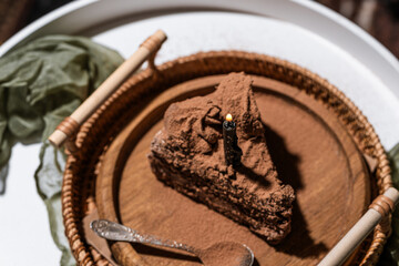 black birthday candle burning on chocolate cake with cream, chocolate cake on a wooden plate and wicker tray on a white coffee table, dark background, birthday cake, flatlay