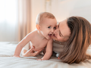 Mother kissing her happy baby on the bed. Loving mother kisses her joyful baby while playing on the bed, sharing a tender and affectionate moment at home.