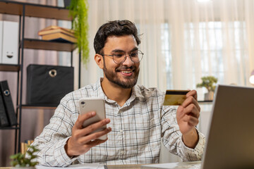 Happy Indian businessman using bank credit card and smartphone for online shopping payments at home...