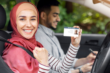Glad muslim woman in hijab show driving license and fist in open door in car after exam with male teacher, outdoor in summer. Winning gesture, get license, driving test in city, lessons in traffic