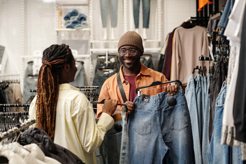 Happy young man holding hanger with blue jeans and looking at his girlfriend helping him choose new attire