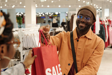 Modern consumer in casualwear passing paperbag to female clerk of showroom while buying new attire...