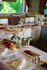 Elegantly set dining table with white tablecloths, white plates, silver cutlery, neatly folded white napkins in napkin rings, and multiple wine glasses at each setting