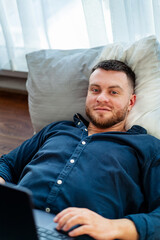 Fototapeta premium Man using laptop while reclining on bed. A man relaxes on his bed, engaging with his laptop while enjoying a peaceful moment indoors.