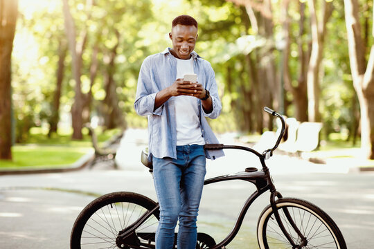 Funky black guy with bicycle sending communicating online on smartphone at park. Handsome African American man checking mail, browsing web, sending message or posting in social media outside - Powered by Adobe