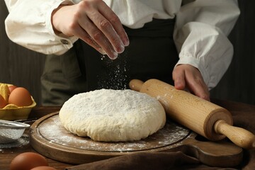 Woman with rolling pin adding flour onto dough at wooden table, closeup