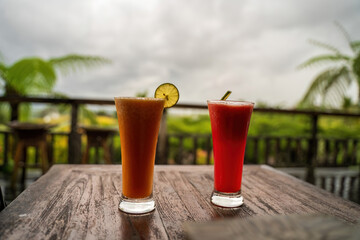 Two refreshing tropical drinks, one orange with a lime slice and one red with a straw, sit on a wooden table with a lush green background in Bali, creating a perfect vacation vibe