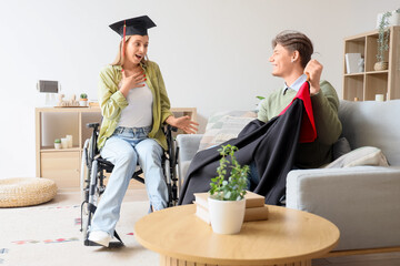 Female graduate in wheelchair and her friend with robe at home