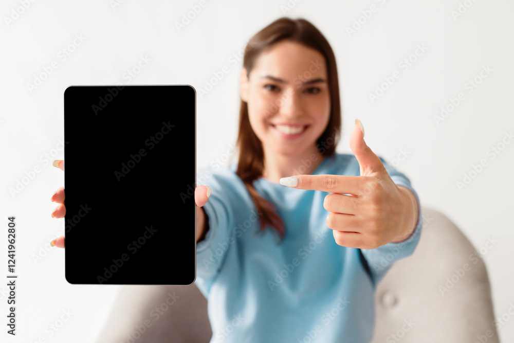 Poster joyful millennial woman pointing at tablet with blank screen for mockup, promoting new app, offering