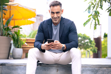 Obraz premium Happy middle-aged man in a blazer sitting on steps while using his smartphone. He has a relaxed and focused expression. The background features an outdoor urban environment with greenery and seating. 