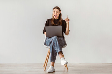 Full length of young Caucasian lady with laptop sitting on chair, pointing upwards, showing IDEA gesture against white wall. Millennial woman having aha moment, feeling excited during online work