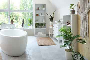 Bathtub, shelving units and houseplants near window in modern bathroom